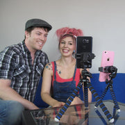 A smiling man and woman sit on a blue couch, recording or livestreaming with two smartphones held by 11 Adjustable Tripod Mounts with Phone Holders atop a glass table.