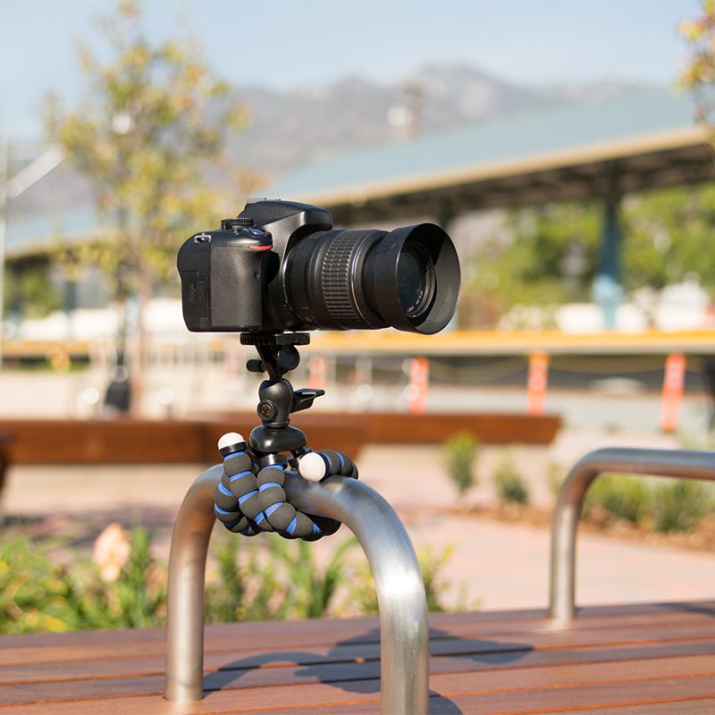 The 11 Inch Flexible Camera Tripod for Canon, Nikon, Samsung, and other 1/4-20 digital cameras is wrapped around an outdoor metal rail, holding a DSLR camera with benches, greenery, and blurred buildings in the background.