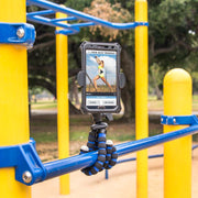 The 11 inch Tripod with RoadVise® Phone Holder Mount for Video is used outdoors on blue playground bars to hold a smartphone displaying a workout video, with trees and greenery in the background.