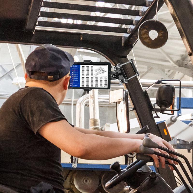 A worker in an industrial facility operates machinery indoors, securely checking a digital screen mounted on the RoadVise® Ultra Forklift Front Guard Phone and Tablet Mount.
