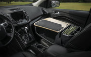 The front seat of the car is transformed into a mobile office using the Arkon Mounts AutoExec GripMaster Efficiency Car Desk, which features a wooden desk surface, a felt mat, and an organizer for file folders. Parked near a grassy field visible through the windows, this passenger seat desk offers convenient workspace on-the-go.