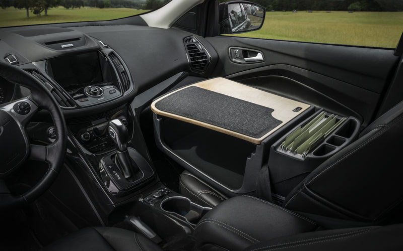 The front seat of the car is transformed into a mobile office using the Arkon Mounts AutoExec GripMaster Efficiency Car Desk, which features a wooden desk surface, a felt mat, and an organizer for file folders. Parked near a grassy field visible through the windows, this passenger seat desk offers convenient workspace on-the-go.