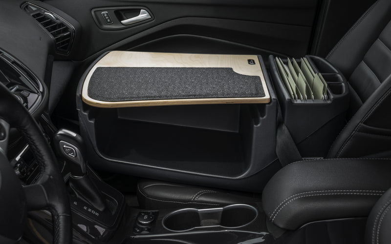 A car interior features the Arkon Mounts AutoExec GripMaster Efficiency Car Desk, equipped with a felt mat and file folder organizer in place of the standard passenger seat, positioned beside the driver’s seat and center console to provide a practical mobile workstation.