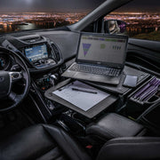 The Arkon Mounts AutoExec GripMaster Car Desk is set up inside a vehicle with a laptop, notepad, pens, and a folder organizer, creating the perfect mobile workstation as the city lights shine through the windows at night.