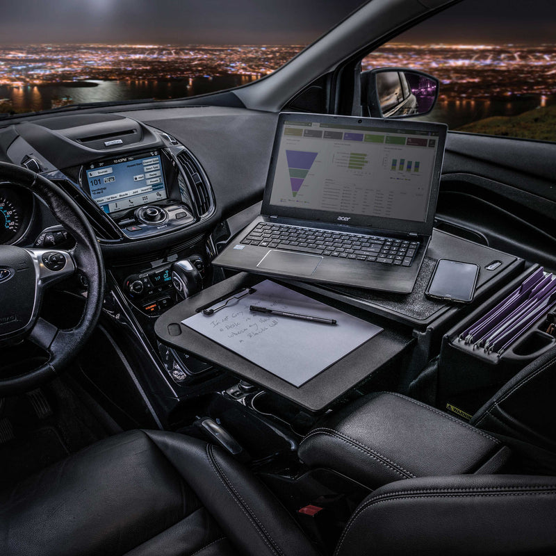 The Arkon Mounts AutoExec GripMaster Car Desk is set up inside a vehicle with a laptop, notepad, pens, and a folder organizer, creating the perfect mobile workstation as the city lights shine through the windows at night.