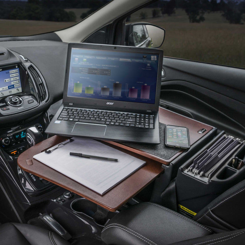 A laptop, phone, notepad, and pen are organized on the AutoExec GripMaster Car Desk by Arkon Mounts, forming an efficient mobile workstation secured to the car’s front passenger seat. The car interior and dashboard are visible, with a scenic view outside the window.