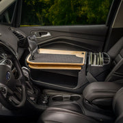 A car interior featuring the AutoExec GripMaster Car Desk with Mega Grip™ Drill-Base Mount attached to the passenger seat, complete with a desk mat and document organizer next to the seat, set against a backdrop of green trees seen through the window.