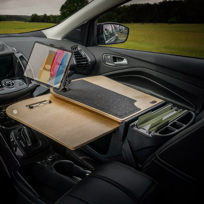 The AutoExec GripMaster Car Desk with Slim-Grip® Ultra Pro Multi-Angle Mount by Arkon Mounts is installed in the front passenger seat of a car parked beside a grassy field, offering a versatile wooden mobile workstation complete with a tablet holder, mouse pad, pen slot, and file holder.