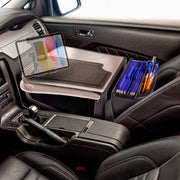 The car interior features an AutoExec GripMaster Car Desk with Slim-Grip® Ultra Pro Multi-Angle Mount by Arkon Mounts, securely mounted on the passenger seat. The desk holds a laptop, blue folders, pens, and a mouse pad—creating an ideal mobile office setup. Additional space accommodates accessories such as a tablet holder. The gear shift and black leather seats are also visible in the scene.
