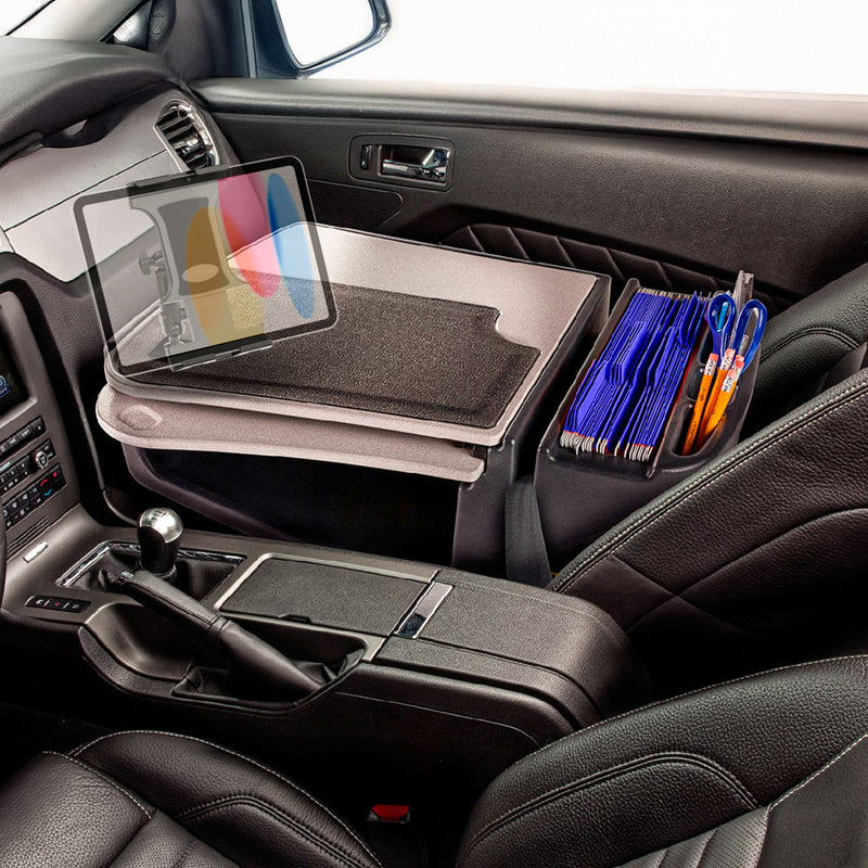 The car interior features an AutoExec GripMaster Car Desk with Slim-Grip® Ultra Pro Multi-Angle Mount by Arkon Mounts, securely mounted on the passenger seat. The desk holds a laptop, blue folders, pens, and a mouse pad—creating an ideal mobile office setup. Additional space accommodates accessories such as a tablet holder. The gear shift and black leather seats are also visible in the scene.
