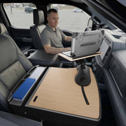 A man sits in a parked vehicle using an Arkon Mounts rugged laptop, working on an AutoExec Reach Front Seat Car Desk that spans the center console and features a large wooden work surface and an organized tray with folders—creating an efficient mobile workspace.