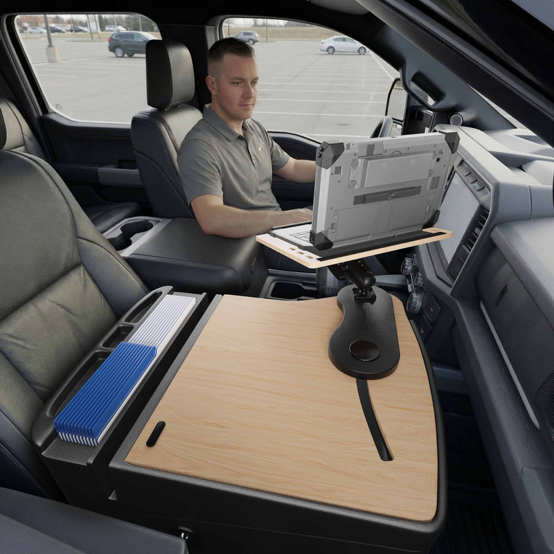A man sits in a parked vehicle using an Arkon Mounts rugged laptop, working on an AutoExec Reach Front Seat Car Desk that spans the center console and features a large wooden work surface and an organized tray with folders—creating an efficient mobile workspace.