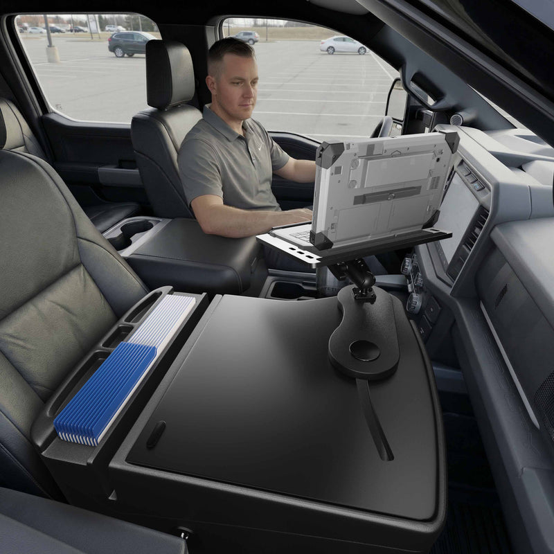 A man sits in the driver’s seat of a parked car, working on his laptop that’s securely mounted to an Arkon Mounts AutoExec Reach Front Seat Car Desk. The front passenger seat and car interior, including a spacious console organizer with neatly arranged files, are visible—showcasing a highly organized mobile workspace.
