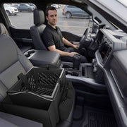 A person in a black shirt sits in the driver seat of a truck, transforming the front cab into a mobile workspace. The Portable Crate Workstation with Apron Organizer – Choose Your Mounting Setup is positioned on the passenger seat, while a travel mug rests securely in the center console cupholder. The vehicle is parked in a lot, fully equipped for productivity on the go.