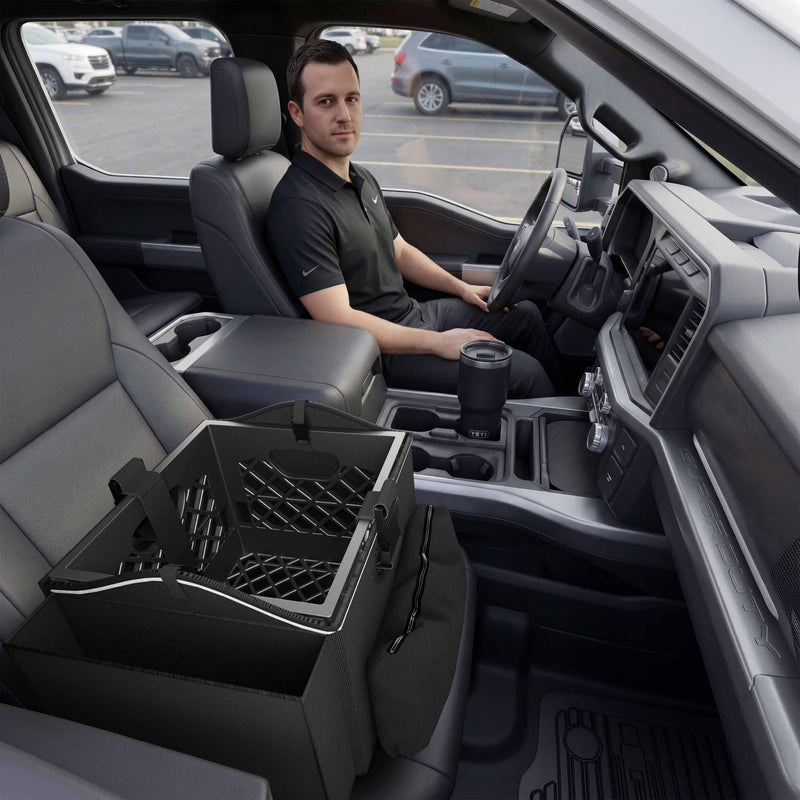 A person in a black shirt sits in the driver seat of a truck, transforming the front cab into a mobile workspace. The Portable Crate Workstation with Apron Organizer – Choose Your Mounting Setup is positioned on the passenger seat, while a travel mug rests securely in the center console cupholder. The vehicle is parked in a lot, fully equipped for productivity on the go.