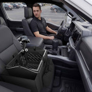 A man sits in the driver seat of a car, looking at the Portable Crate Workstation with Apron Organizer – Choose Your Mounting Setup, which features partitions and a mounted device on the passenger seat. The car’s interior is modern, and a parking lot is visible outside.