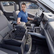 A man in a blue shirt sits in a parked car, reaching toward his phone mounted on the Portable Crate Workstation with Apron Organizer – Choose Your Mounting Setup, which is attached to a black crate on the passenger seat, creating an efficient mobile workstation. The car interior is modern and clean.