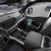 The interior of a parked pickup truck has been converted into a mobile workspace, featuring a tablet secured in a device mounting system, a cup resting in the cupholder, and the Portable Crate Workstation with Apron Organizer – Choose Your Mounting Setup neatly arranged on the front passenger seat; other vehicles are visible through the window in the parking lot.
