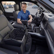 A man in a blue polo shirt sits in the driver’s seat of a parked truck, which serves as his mobile workstation. A mounted tablet and the Portable Crate Workstation with Apron Organizer – Choose Your Mounting Setup rest on the passenger seat, while a silver tumbler sits in the center console.