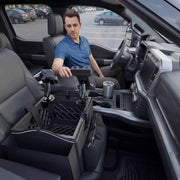A man sits in the driver’s seat of a modern parked car, using the Portable Crate Workstation with Apron Organizer—featuring his chosen mounting setup—in the front passenger area, with a cup and phone visible nearby.