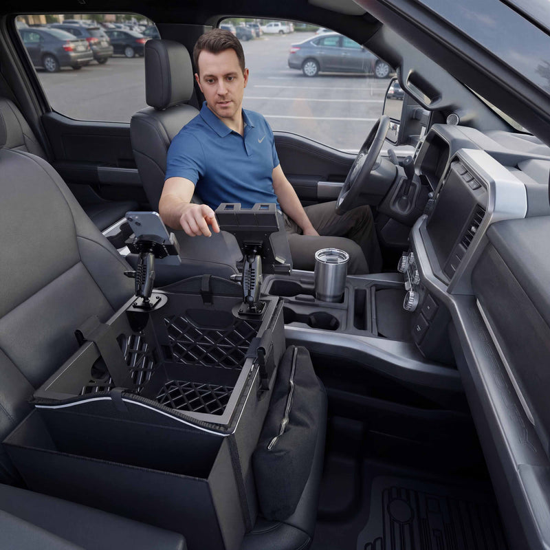 A man sits in the driver’s seat of a modern parked car, using the Portable Crate Workstation with Apron Organizer—featuring his chosen mounting setup—in the front passenger area, with a cup and phone visible nearby.