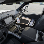 The interior of a vehicle features a GripMaster Efficiency Car Desk with Mega Grip™ Drill-Base Mount, providing a spacious flat workspace over the center console. A phone is securely mounted on the integrated drill-base mount, while a travel mug rests nearby. Through the windows, an empty parking lot is visible—creating an ideal mobile workstation for productivity on the go.
