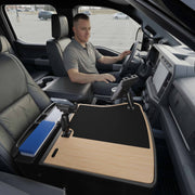 A man sits in the driver’s seat of a parked truck, utilizing the AutoExec GripMaster Efficiency Car Desk with Mega Grip™ and Slim-Grip® Ultra Pro Drill-Base Mounts—a large wooden work surface with office supplies arranged across the front passenger seat area, conveniently featuring a tablet holder for added efficiency.
