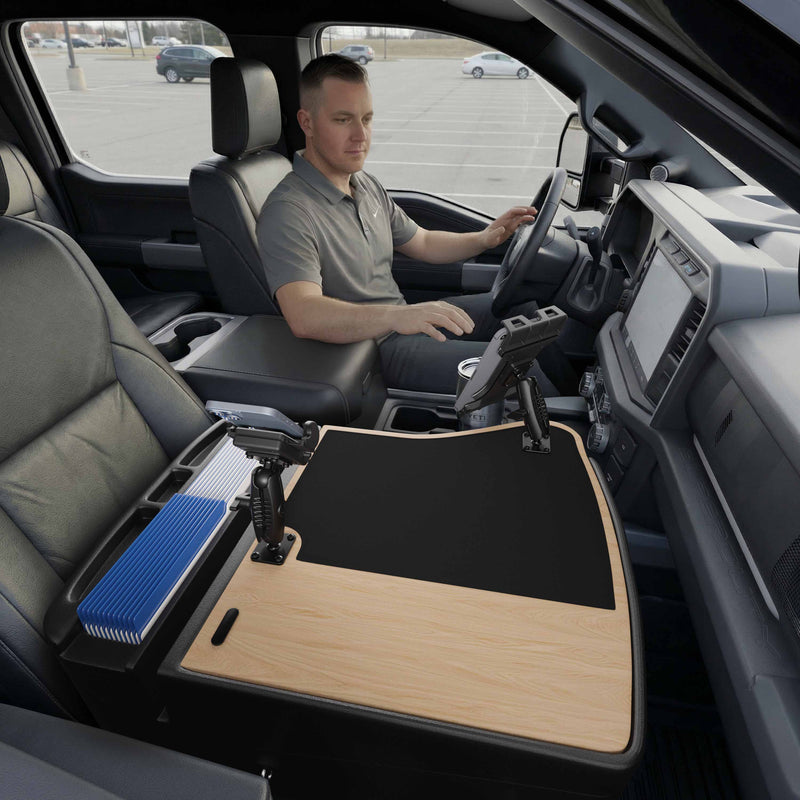 A man sits in the driver’s seat of a parked truck, utilizing the AutoExec GripMaster Efficiency Car Desk with Mega Grip™ and Slim-Grip® Ultra Pro Drill-Base Mounts—a large wooden work surface with office supplies arranged across the front passenger seat area, conveniently featuring a tablet holder for added efficiency.