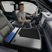 A man sits in the driver’s seat of a parked vehicle, creating an efficient mobile workstation with the GripMaster Efficiency Car Desk featuring a Mega Grip™ Drill-Base Mount—he holds folders and uses a mounted device positioned over the center console, while the passenger seat beside him remains empty.