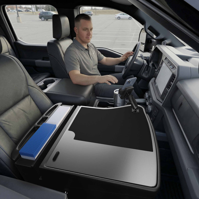 A man sits in the driver’s seat of a parked vehicle, creating an efficient mobile workstation with the GripMaster Efficiency Car Desk featuring a Mega Grip™ Drill-Base Mount—he holds folders and uses a mounted device positioned over the center console, while the passenger seat beside him remains empty.