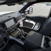 The front seat interior of a modern pickup truck features the GripMaster Efficiency Car Desk with Mega Grip™ Drill-Base Mount securing a phone, alongside a stainless steel travel mug, large touchscreen display, and a fold-out workstation on the passenger side. The truck is parked in an empty lot.