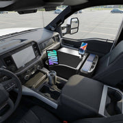 The interior of a truck features the AutoExec GripMaster Efficiency Car Desk, equipped with Mega Grip™ and Slim-Grip® Ultra Pro Drill-Base Mounts, securely holding a tablet and smartphone next to a stainless steel travel mug on the workspace in the center console, while parked in an empty lot.
