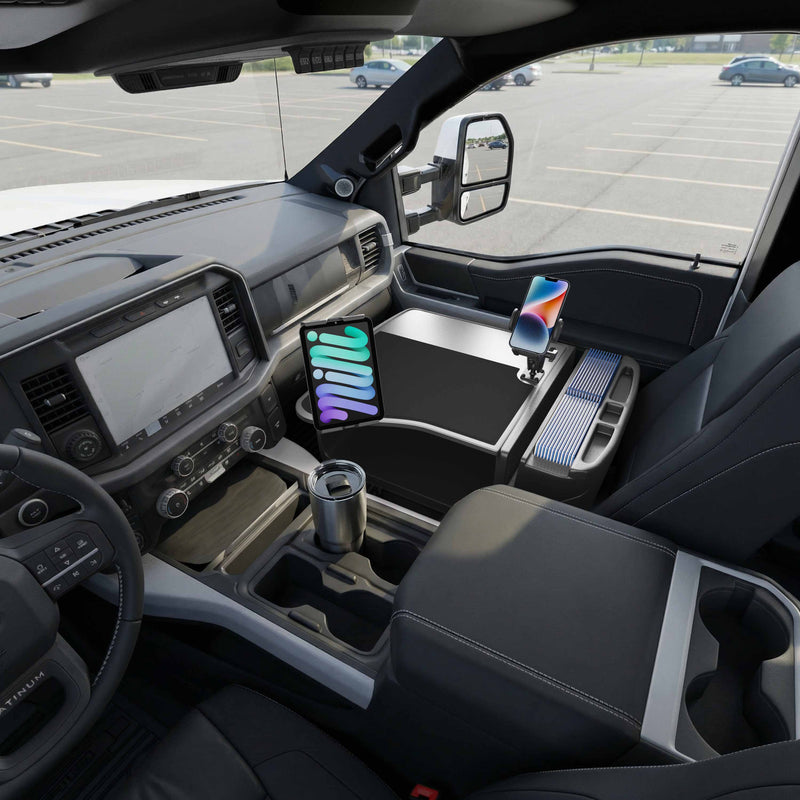 The interior of a truck features the AutoExec GripMaster Efficiency Car Desk, equipped with Mega Grip™ and Slim-Grip® Ultra Pro Drill-Base Mounts, securely holding a tablet and smartphone next to a stainless steel travel mug on the workspace in the center console, while parked in an empty lot.