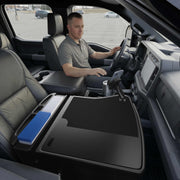 A man sits in the driver’s seat of a parked truck. The front passenger seat features the GripMaster Efficiency Car Desk with Mega Grip™ Drill-Base Mount, creating a flat mobile workstation with neatly organized folders and a secure mounted phone holder.