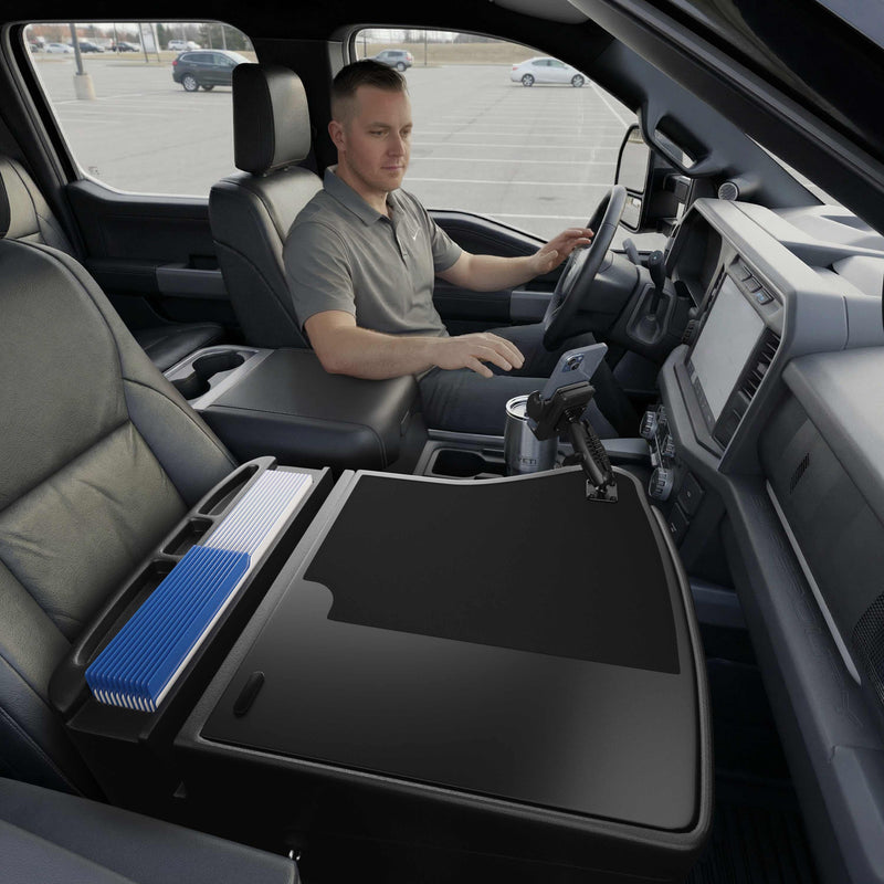 A man sits in the driver’s seat of a parked truck. The front passenger seat features the GripMaster Efficiency Car Desk with Mega Grip™ Drill-Base Mount, creating a flat mobile workstation with neatly organized folders and a secure mounted phone holder.