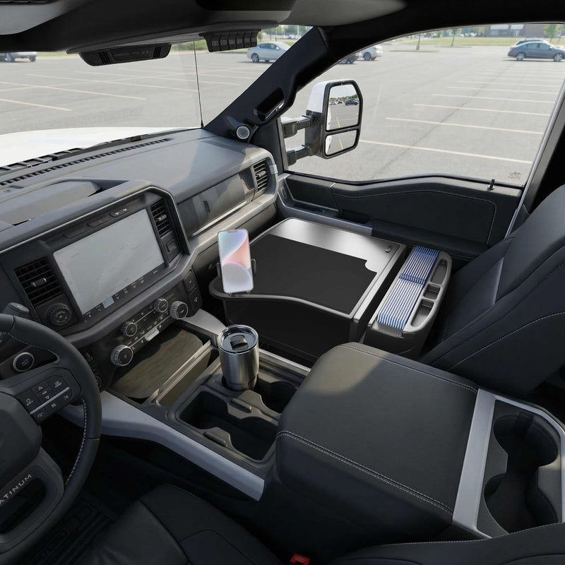 The interior of a modern truck is transformed into a convenient mobile workstation with the GripMaster Efficiency Car Desk featuring a Mega Grip™ Drill-Base Mount. The fold-out work surface supports a mounted phone and stainless steel travel mug, while the dashboard and controls remain clearly visible. A parking lot can be seen through the window, and the sturdy Mega Grip™ Drill-Base Mount keeps essentials securely in place.
