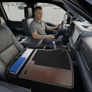 A man sits in the driver’s seat of a parked truck, working at his mobile office set up on the GripMaster Efficiency Car Desk with a stylish wood finish, which is equipped with office supplies and features a Mega Grip™ Drill-Base Mount securely holding his phone in the front passenger area.