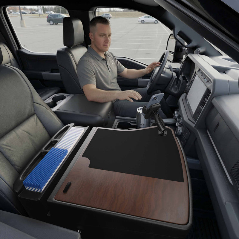 A man sits in the driver’s seat of a parked truck, working at his mobile office set up on the GripMaster Efficiency Car Desk with a stylish wood finish, which is equipped with office supplies and features a Mega Grip™ Drill-Base Mount securely holding his phone in the front passenger area.