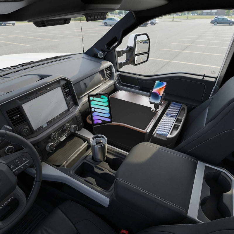 The interior of a parked vehicle showcases the AutoExec GripMaster Efficiency Car Desk, equipped with Mega Grip™ and Slim-Grip® Ultra Pro Drill-Base Mounts securely holding a mounted tablet, as well as a phone holder with a smartphone. A stainless steel travel mug rests beside what appears to be an empty passenger seat. The convenient car desk and professional-grade mounts enhance the mobile workspace, with the dashboard and controls clearly visible.