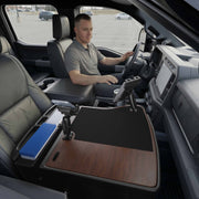 A man sits in the driver’s seat of a parked truck, utilizing the AutoExec GripMaster Efficiency Car Desk with Mega Grip™ and Slim-Grip® Ultra Pro Drill-Base Mounts over the center console. The fold-out desk features a wood surface, black mat, pen holders, a blue file organizer, and an integrated tablet holder for added convenience while working on the road.