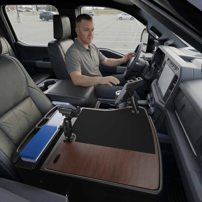 A man sits in the driver’s seat of a parked truck, utilizing the AutoExec GripMaster Efficiency Car Desk with Mega Grip™ and Slim-Grip® Ultra Pro Drill-Base Mounts over the center console. The fold-out desk features a wood surface, black mat, pen holders, a blue file organizer, and an integrated tablet holder for added convenience while working on the road.