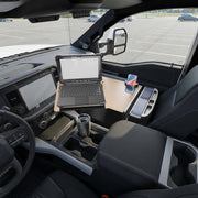 A laptop sits atop the AutoExec Reach Front Seat Car Desk with RoadVise® Ultra Pro Drill-Base Mount, which is securely attached to the car’s center console to create an efficient mobile workstation. A smartphone rests in the dedicated RoadVise® phone holder beside a travel mug, while an empty parking lot is visible through the windshield.