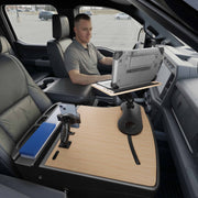 A man sits in the driver’s seat of a parked vehicle, using a laptop mounted on the AutoExec Reach Front Seat Car Desk with RoadVise® Ultra Pro Drill-Base Mount—a wooden workstation setup that covers the center console and extends over the passenger seat, creating an efficient mobile workstation.