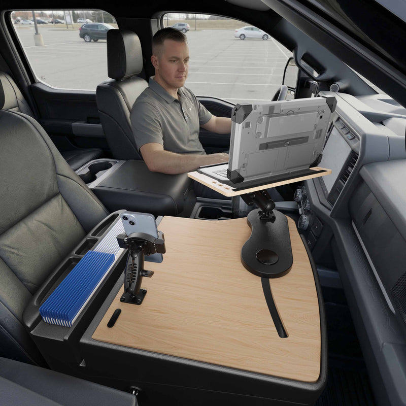 A man sits in the driver’s seat of a parked vehicle, using a laptop mounted on the AutoExec Reach Front Seat Car Desk with RoadVise® Ultra Pro Drill-Base Mount—a wooden workstation setup that covers the center console and extends over the passenger seat, creating an efficient mobile workstation.