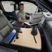 A man sits in a parked vehicle, using a laptop mounted on the AutoExec Reach Front Seat Car Desk by Arkon Mounts, which features the Slim-Grip® Ultra Pro Drill-Base Mount for added stability. A tablet holder and folders are also neatly organized on the desk, creating a functional mobile workspace.