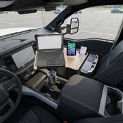 The AutoExec Reach Front Seat Car Desk with Slim-Grip® Ultra Pro and RoadVise® Ultra Pro Drill-Base Mount is set up inside a parked vehicle, holding a laptop, a tablet secured in the included holder, a mug, and various small items, with an empty parking lot visible through the driver’s side window.