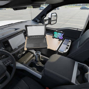 The AutoExec Reach Front Seat Car Desk with LockVise™ Locking Tablet Multi-Angle Mount is set up in the front seat of a parked vehicle, supporting both a laptop and a tablet for an efficient mobile workstation. A stainless steel travel mug rests in the integrated cup holder, with the parking lot visible through the windows.