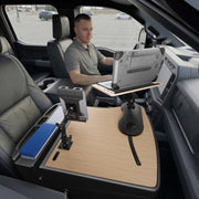 A man sits in a parked vehicle using the AutoExec Reach Front Seat Car Desk with Slim-Grip® Ultra Pro Multi-Angle Mount by Arkon Mounts, featuring a wooden car desk and multi-angle laptop and tablet holders attached to the passenger seat area.