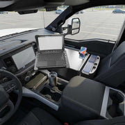 An AutoExec Reach Front Seat Car Desk with RoadVise® Ultra Pro Drill-Base Mount supports a rugged laptop, smartphone, and travel mug, transforming the vehicle’s front seat into a fully equipped mobile workstation; the integrated phone holder securely cradles the device as empty parking spaces stretch out beyond the window.
