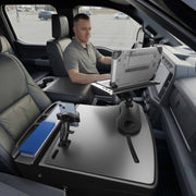 A man sits in the driver’s seat of a parked vehicle, working on his laptop mounted on the AutoExec Reach Front Seat Car Desk with RoadVise® Ultra Pro Drill-Base Mount. His mobile workstation stretches across the front seats and includes a phone holder, blue file organizer, and various accessories.